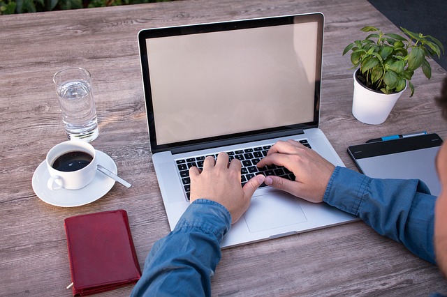 člověk u počítače, notebook na dřevěném stole, vedle káva, voda a zelená kytka